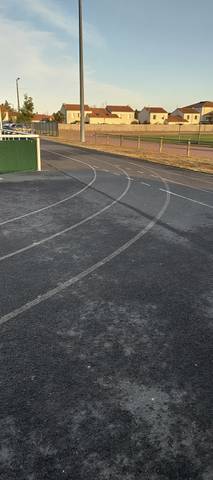 Le virage : enrobé sombre et lignes blanches délimitant les deux couloirs de l'anneau, pavillons du bourg en fond — Complexe Sportif Hugues Martin, La Chevrolière