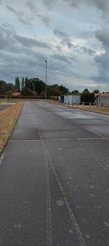 La ligne droite : enrobé gris, joints longitudinaux ouverts entre les bandes, mousse dans les fissures et lignes de couloir pâlies — Complexe Sportif Gauvrit, Sainte-Pazanne
