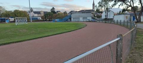 Le virage en fin de journée : cendrée rouge régulière et sans couloir tracé, terrain de football en gazon au centre, clôture bois puis club-house et clocher du bourg en fond — Stade Rene Masse, Saint-Sébastien-sur-Loire