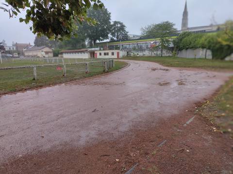 Le virage un jour de pluie : cendrée rouge saturée d'eau, flaques sur toute la largeur, clôture de la pelouse avec son panneau d'interdiction et vieux bâtiment de vestiaires à portes rouges au fond — Complexe Vertou Centre, Vertou