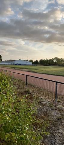 La ligne droite depuis le bord ouest, à travers le grillage : cendrée rougeâtre sans marquage de couloir visible, bâtiments et mâts d'éclairage du complexe au fond — Complexe sportif de la Cailletière, Challans