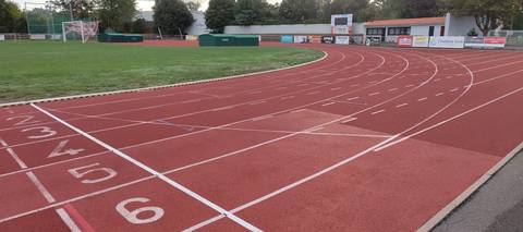 Les couloirs numérotés au sol à l'entrée du virage : tartan rouge, bordure et caniveau contre la pelouse, panneaux des partenaires et tableau d'affichage au fond — Complexe sportif Jean Léveillé, Challans