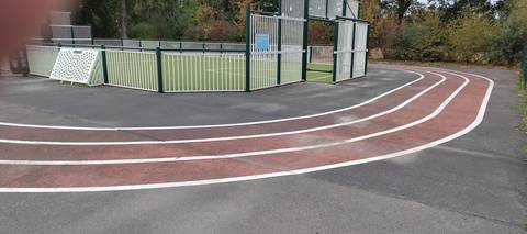 Le virage : trois couloirs en enrobé rouge lignés de blanc, peints sur la dalle d'enrobé gris qui entoure le city-park clôturé et son terrain en gazon synthétique — Complexe sportif des Grenais, Saint-Philbert-de-Grand-Lieu