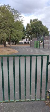 La barrière métallique verte fermée, à la peinture écaillée, qui barre l'entrée ; derrière elle le tracé en enrobé sombre et ses trois couloirs marqués en blanc, un module de jeux pour enfants et un bâtiment modulaire clair — Piste de La Chevrolière, La Chevrolière